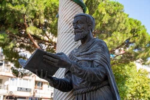 A metal statue of Galen holding a book in front of a pillar, some trees and an apartment block