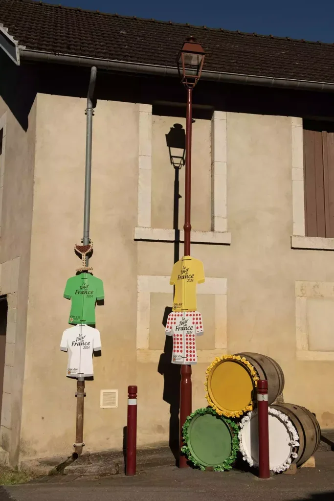 A gutter and lamppost in front of a stone house decorated with  four cardboard signs in the shape of Tour de France jerseys, one in white, one in green, one in yellow and one with red spots on white and a stack of three barrels with the ends painted in yellow, green and white 