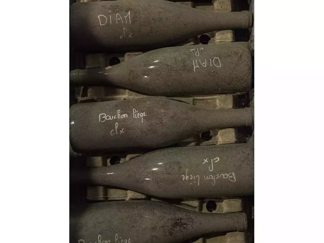 Dusty dark bottles in a cellar with chalk words indicating diam and cork written on the side.