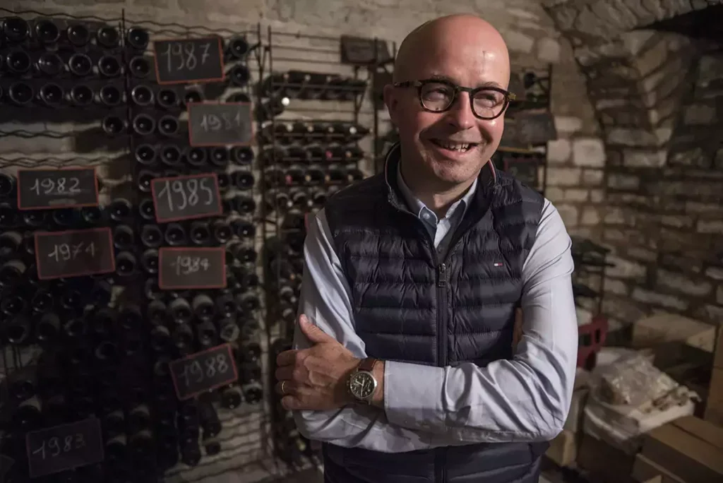 Laurent Delauney, a man in glasses and gilet, stands before some bottles in a cellar laughing