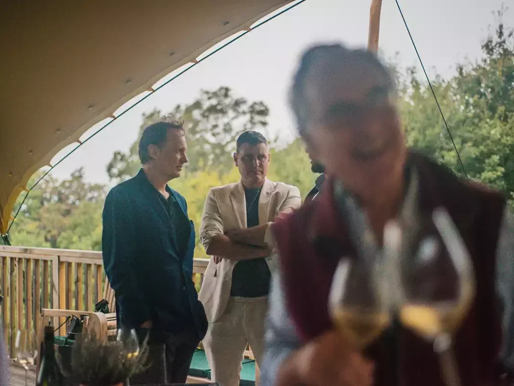 Nick Clarke and Benjamin Abric standing in the background under a marquee at Weyborne Estate with a blurred man, glass and bottle in the foreground