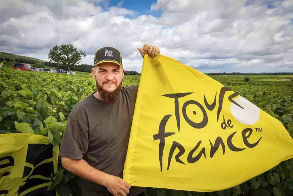 A man in a black t-shirt and a black cap with the word "Jura" on the front holds a yellow Tour de France flag in a vineyard.