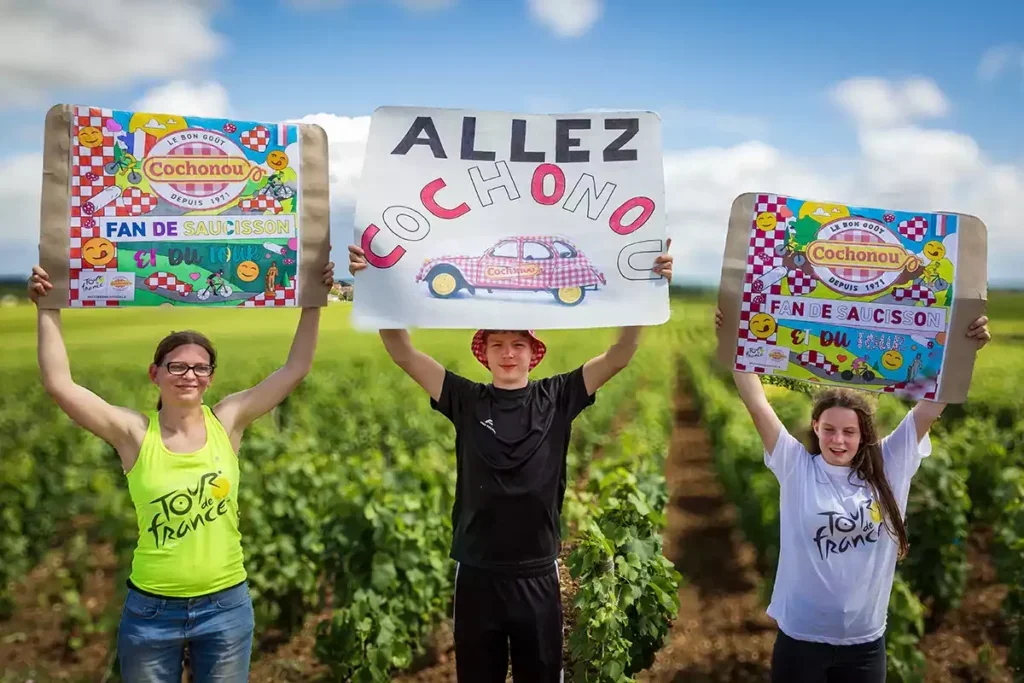 A mother in yellow-green vest and glasses, her son in black shorts and t-shirt and her daughter in white Tour de France t-shirt stand in a line in the vineyard each holding up a homemade signs in support of the Cochounou sausage company, one of the event's sponsors