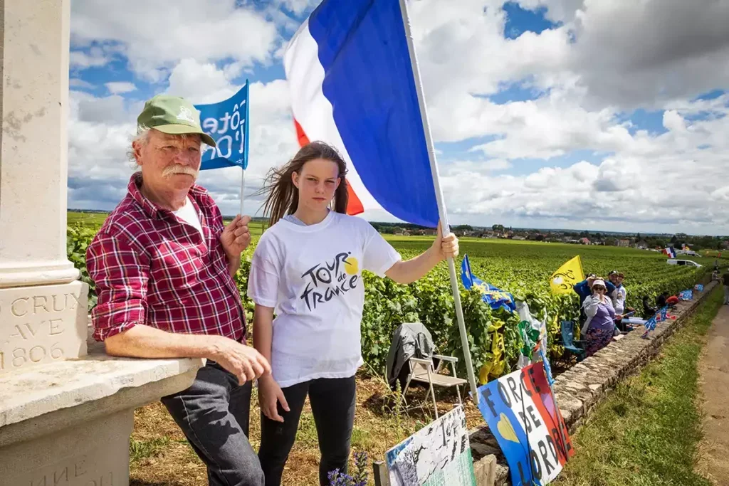 A man with a white moustache and a green cap and red checked shirt stands holding a flag while leaning on a stone pillar in a vineyard in Burgundy with his daughter in a white Tour de France T-shirt carrying a large French flag