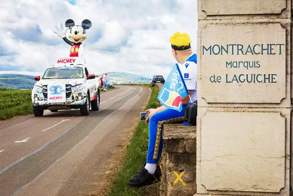 A flatbed truck with a large inflatable figure of Mickey Mouse passes a stone sign for Montrachet Marquis de Laguiche and a man with yellow hair sits on a wall watching