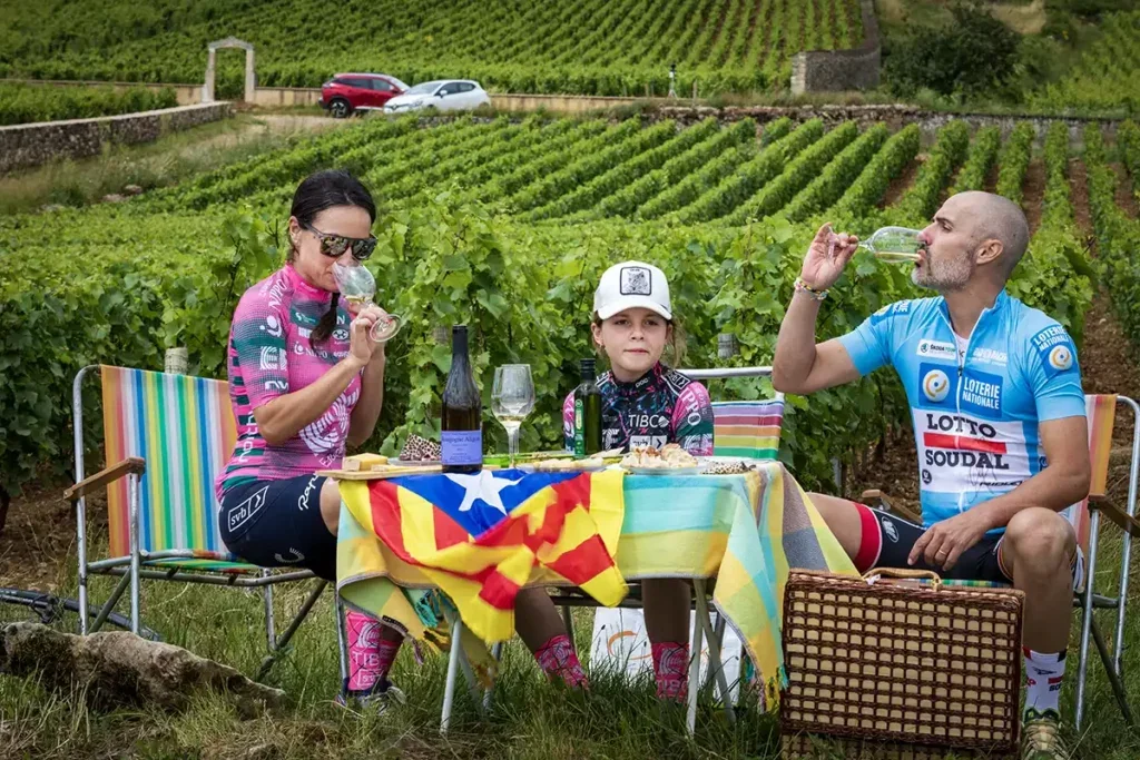 A woman a young girl and a man in cycling clothes sit at a picnic table covered in a Catalan flag in a vineyard drinking white wine. 
